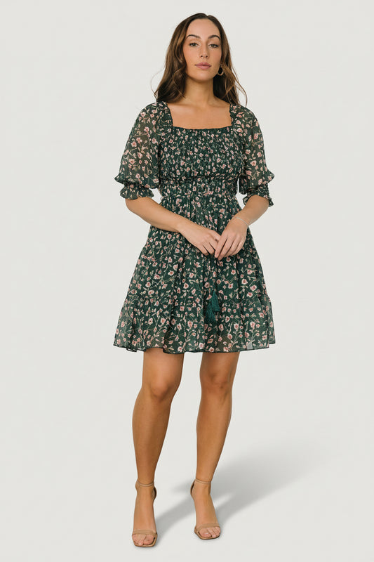 Front view of green and pink floral smocked short dress on a model, showing smocked bodice, ruffled square neckline and tiered short skirt for movement.