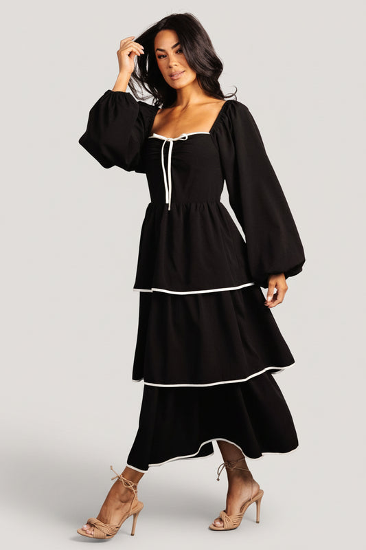Front view of black tiered midi dress on model, showing soft sweetheart neckline and decorative bow at center bust, full-length.