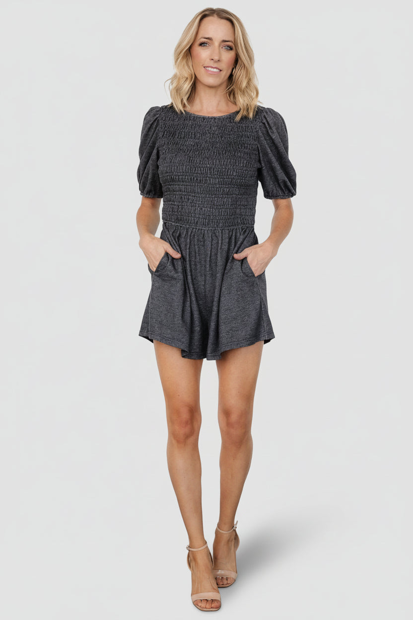 Front view of Beaumont Romper in heather black showing smocked bodice, round neckline, mid-length sleeves, elastic cuffs, and side pockets for daytime wear.