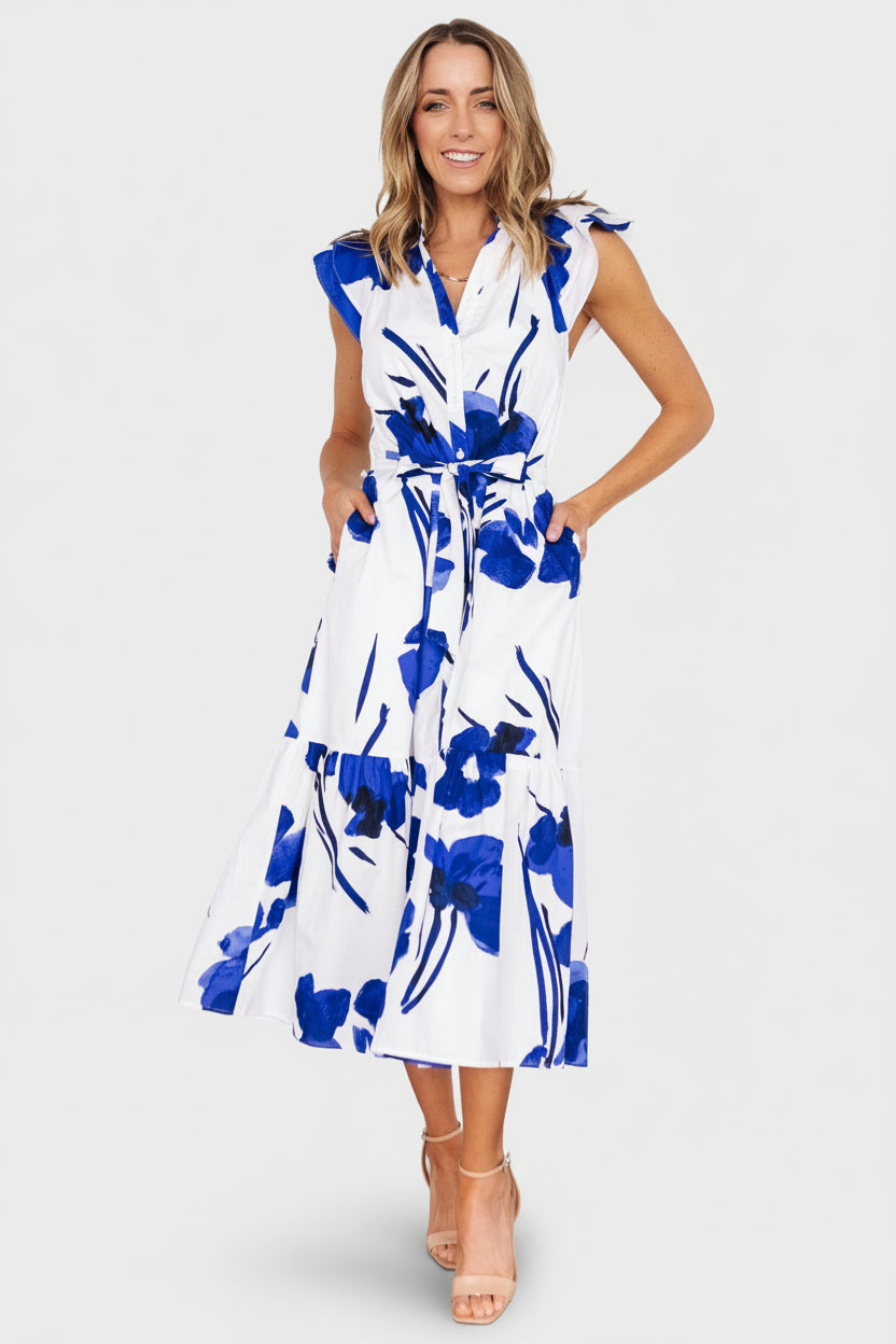 Front view of a woman wearing a white midi dress with cobalt floral print, ruffled notched neckline and button-front, hands in pockets.