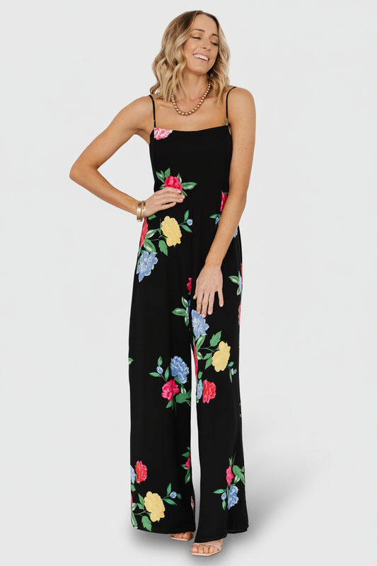 Front view of black floral wide-leg jumpsuit with square neckline, adjustable spaghetti straps, flowing chiffon fabric and full lining for coverage.