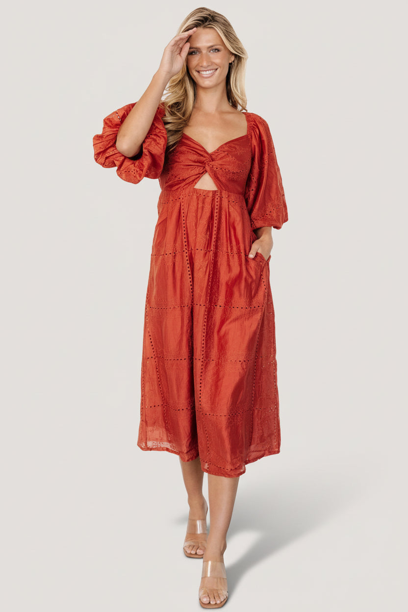 Model wearing Brynn rust midi dress walking pose showcasing silhouette, puff sleeves, and embroidered finish across the dress.