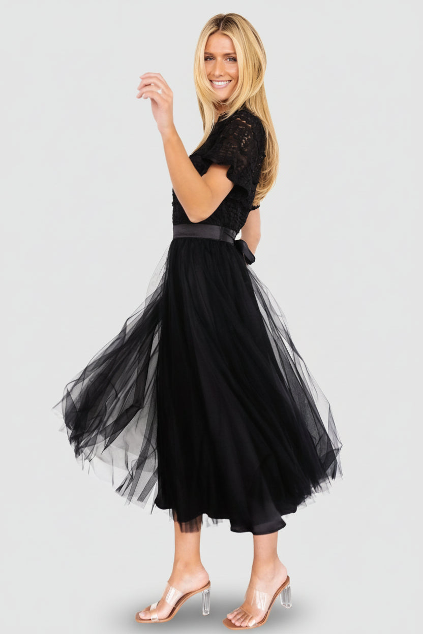Three-quarter studio view showing lace bodice detail, ruffle overlay at the neckline, and the flowing layered tulle skirt of the black midi dress.