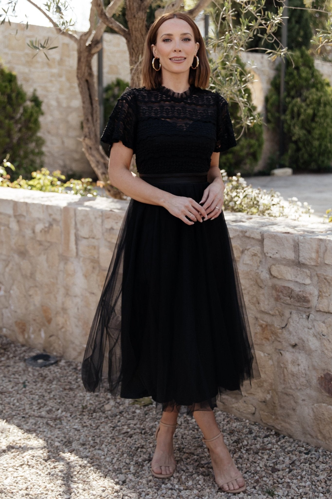 Back view showing the hidden zipper, satin tie at the waist, and the layered tulle skirt falling to a midi length on a neutral backdrop.