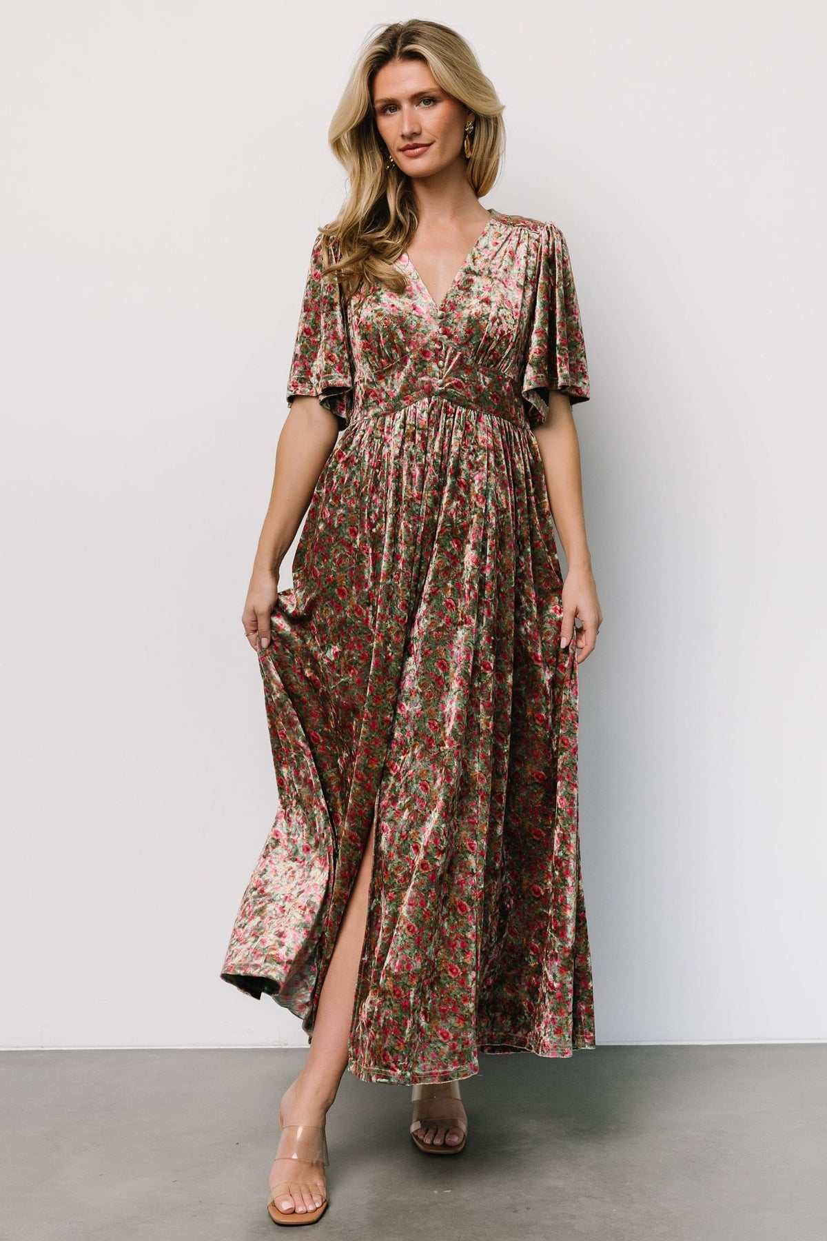 Front view of green multi floral velvet maxi dress on model showing deep V-neck, gathered bust, flutter sleeves and left leg slit for movement.