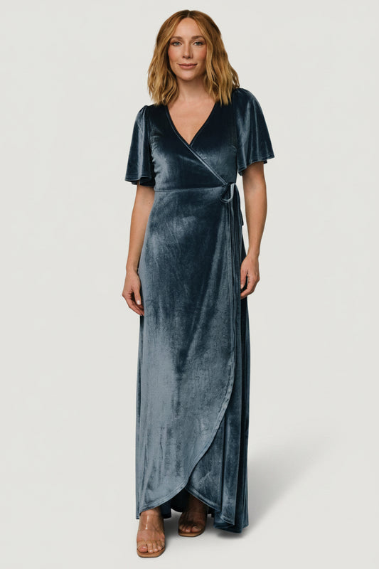 Three-quarter angle of velvet wrap dress in soft blue highlighting tulip-style skirt, drape and fabric texture in motion.