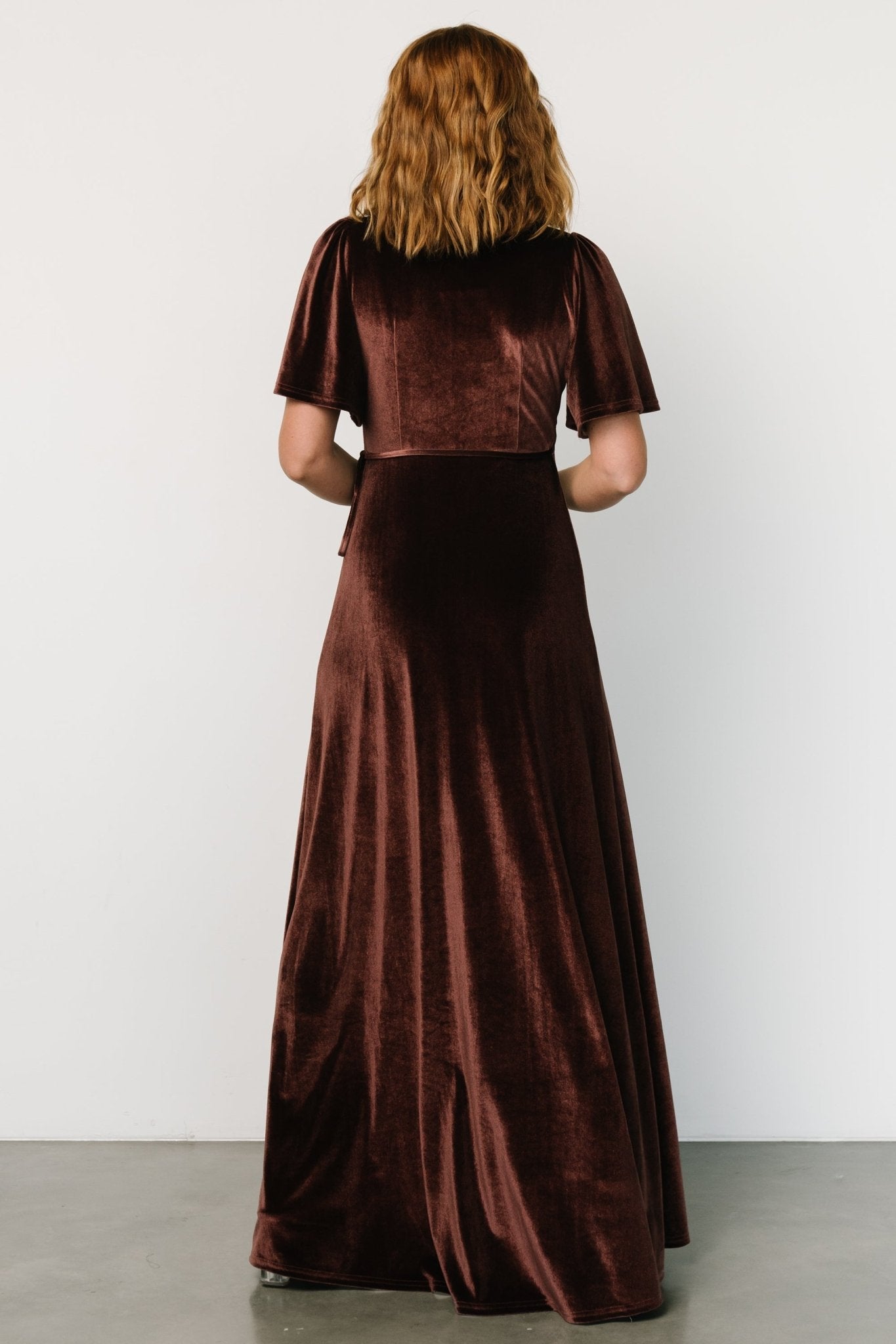 Side view of the clove velvet maxi dress displaying the tulip-style skirt movement, waist tie detail, and medium flutter sleeve silhouette.