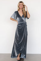 Front view of dusty blue velvet wrap maxi dress on model showing V-neck, flutter sleeves, and tulip skirt with natural drape and movement, fully lined.