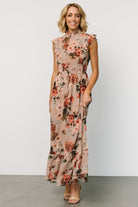 Full-length model shot of blush floral chiffon maxi dress showing the smocked waist, flutter sleeves, and gentle ruffle movement at the hem.