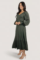 Side profile of the deep olive midi dress showing smocked bodice, elastic shoulder detail, and tiered hem on a model.