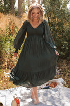 Front view of a woman wearing a deep olive smocked dotted midi dress with V‑neck and long sleeves, studio background.