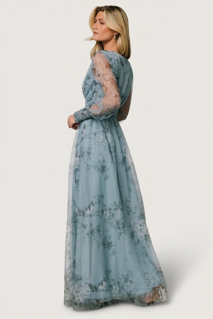 Back and side view of dusty blue floral tulle dress showing sheer long sleeves, raw hem edge, and gentle A-line skirt drape.