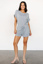 Lely romper in dusty blue — front view showing textured vertical ribs, elastic waist, faux tie, short cuffed sleeves, and front pockets.