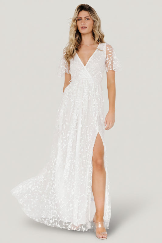Front view of Marseille vine-embossed organza maxi dress in white, showing crossover surplice neckline, elastic waist with tie, left-leg slit and raw cut hemline.