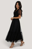Side view of black tulle midi dress highlighting elastic V-neck, flirty short sleeves and the gentle gather of the tiered skirt silhouette.