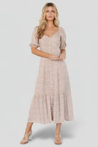 Front view of a beige floral maxi dress with wide scoop neckline, button-front, puffed sleeves and flowing tiered skirt, shown on a model.