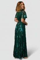 Side profile of Muse Sequin Maxi Dress in emerald green, illustrating fit through the waist, sash detail, and movement of the skirt.
