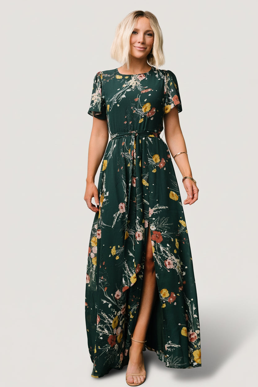 Front view of dark green floral chiffon maxi dress showing rounded neckline, short flutter sleeves, removable waist tie, and left side slit for movement.