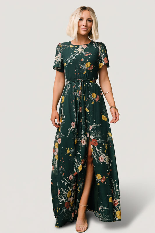 Front view of dark green floral chiffon maxi dress showing rounded neckline, short flutter sleeves, removable waist tie, and left side slit for movement.