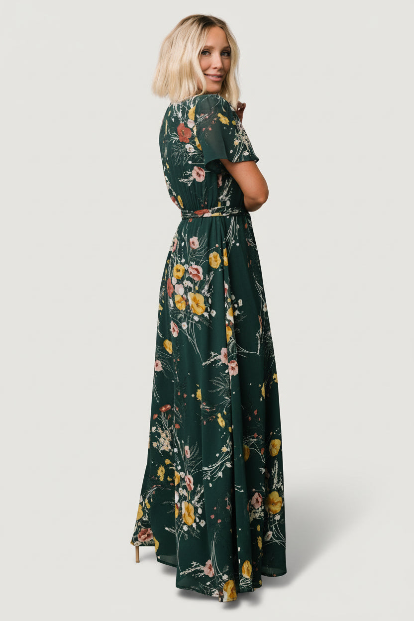Full-length image of model wearing dark green floral maxi dress, illustrating maxi sweep and left side slit while standing.