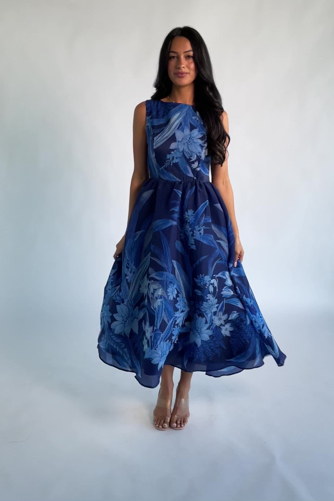 Model wearing a navy sleeveless midi dress with a high boat neckline and a fitted bodice. A tonal blue floral print adds subtle dimension throughout, while the defined waist flows into a full midi skirt that creates graceful movement and an elegant, timeless silhouette.