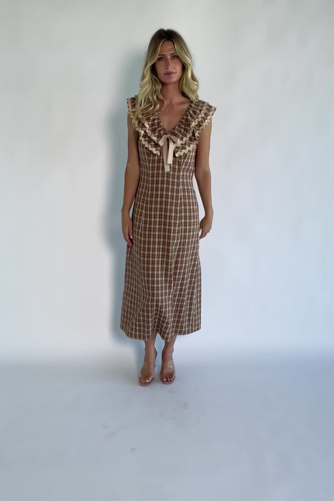 Model wearing a dusty mocha and cream plaid midi dress with a V-neckline, decorative bow tie detail, cascading ruffle trim, and a fitted silhouette.
