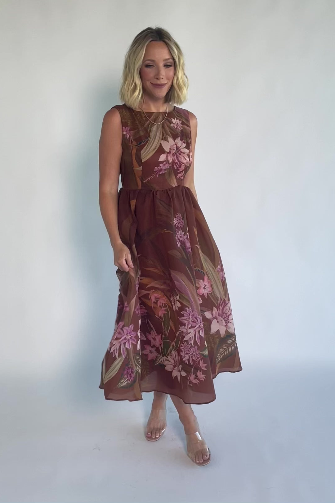 Model wearing a brown floral midi dress with a high boat neckline, sleeveless fitted bodice, and defined waist. The dress features a pink floral print throughout and a flowing midi-length skirt, creating an elegant, timeless silhouette.