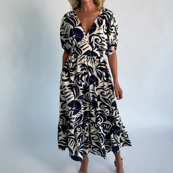 Woman wearing a navy and ivory printed midi dress with a V-neckline, short sleeves, an empire waist, and a tiered hem.
