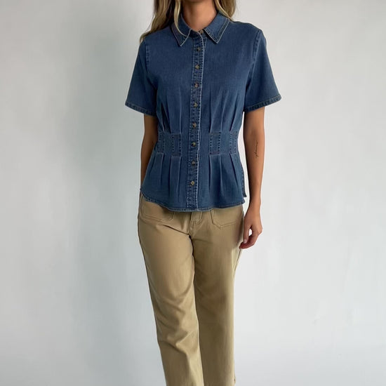 Medium wash stretch denim peplum top with a collared neckline, button-front closure, short sleeves, tailored seam detailing, and a flattering fitted silhouette.