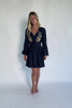 Model wearing a dark chambray short dress with a V-neckline, floral embroidery at the bust, long sleeves with elastic cuffs, and a double elastic waistband.