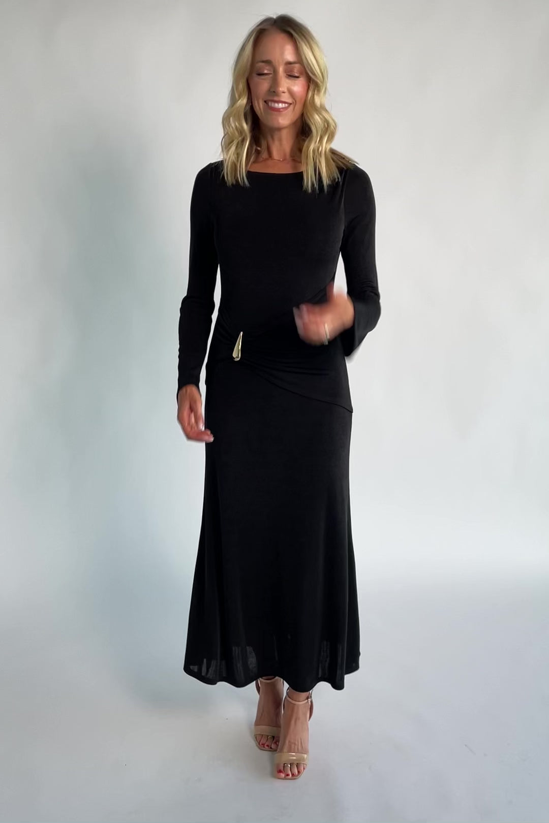 Model wearing a black long-sleeve maxi dress with a fitted silhouette and round neckline. Draped fabric gathers at the waist and is accented by a gold-tone metal detail.
