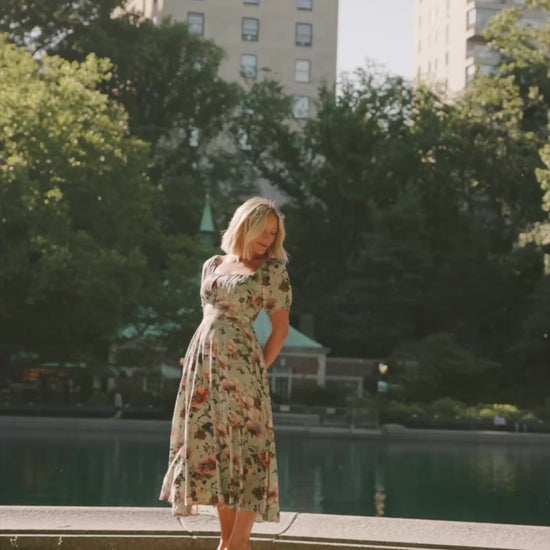 Model wearing the Charlotte Midi Dress in Sage, a sage green midi dress with a pink multi floral print, rounded square neckline, puff sleeves, banded waist, and flowing A-line skirt with side pockets.