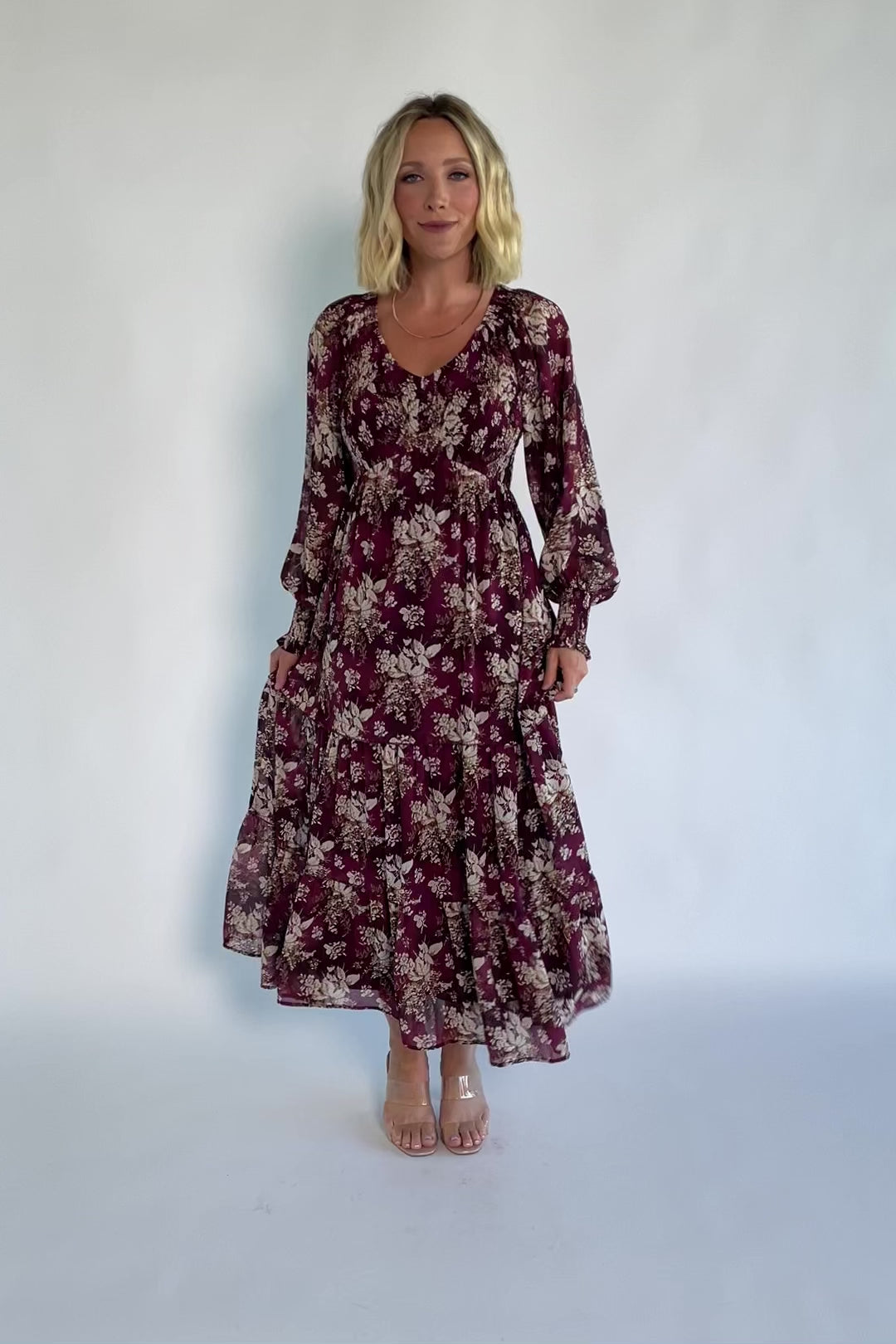 Model wearing a burgundy floral maxi dress with a V-neckline, smocked bodice, and long sheer sleeves with smocked ruffle cuffs.