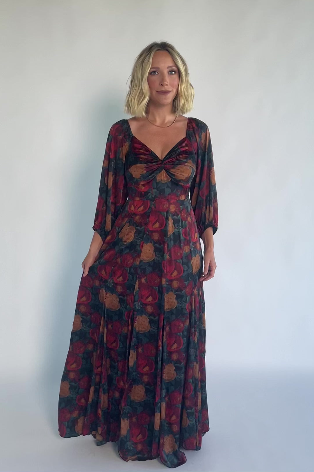 Model wearing a dark jade maxi dress with an oversized marsala floral print, a twist-knot V-neckline, half sleeves with elastic cuffs, a defined high waist, and a flowing maxi skirt with godet panels and an asymmetrical scalloped hem.