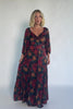 Model wearing a dark jade maxi dress with an oversized marsala floral print, a twist-knot V-neckline, half sleeves with elastic cuffs, a defined high waist, and a flowing maxi skirt with godet panels and an asymmetrical scalloped hem.