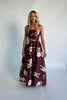 Model wearing a strapless maxi gown with a mahogany floral print and a fitted bodice. A rosette detail accents the neckline, while the flowing floor-length skirt creates elegant movement.