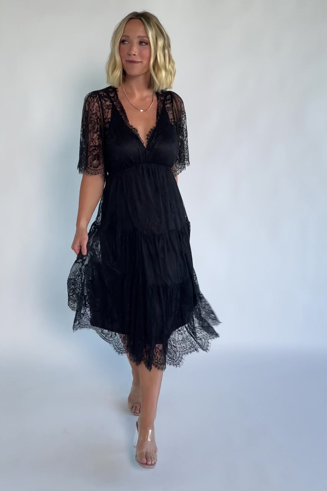 Model wearing a black lace midi dress with a deep V-neckline, short sheer scalloped sleeves, and an elastic waist.