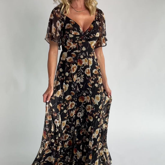 Black floral chiffon maxi dress with a sweetheart neckline, flutter sleeves, smocked back bodice, tiered skirt, and subtle gold foil detailing.