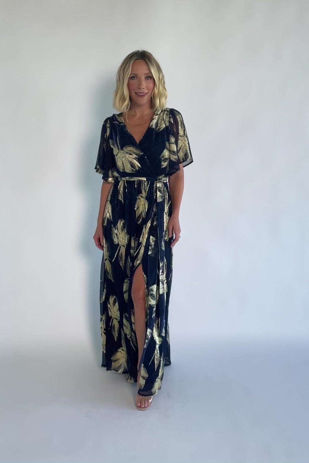 Navy and gold shimmer maxi dress with a surplice V-neckline, sheer sleeves, an elastic waist with a removable sash tie, and a flowing full-length skirt with a side slit.