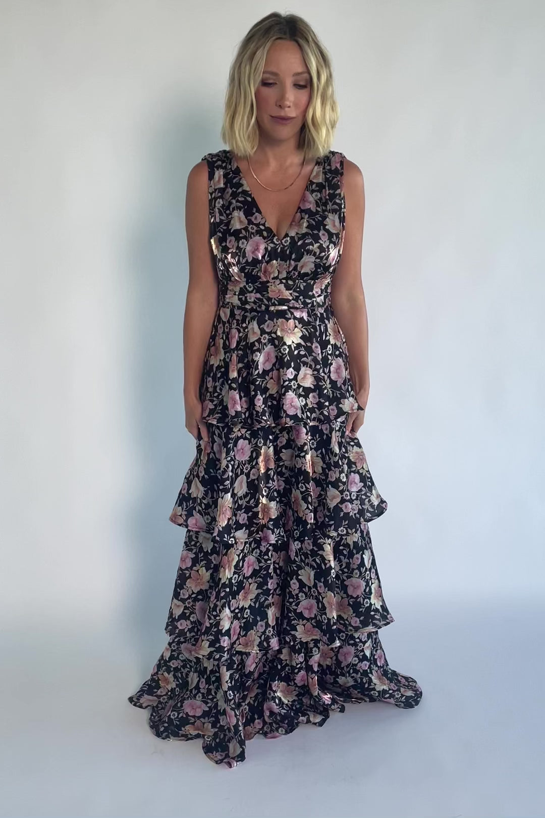 Model wearing a black floral maxi dress with a deep V-neckline, banded waist, and cascading tiered skirt. The dress features a black background with pink, yellow, and orange floral print, creating a flowing, romantic silhouette.