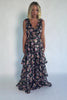 Model wearing a black floral maxi dress with a deep V-neckline, banded waist, and cascading tiered skirt. The dress features a black background with pink, yellow, and orange floral print, creating a flowing, romantic silhouette.