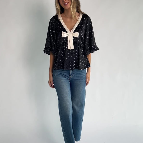 Black satin polka dot peplum top with an ivory contrast collar, decorative bow, V-neckline, and elbow-length ruffle sleeves.