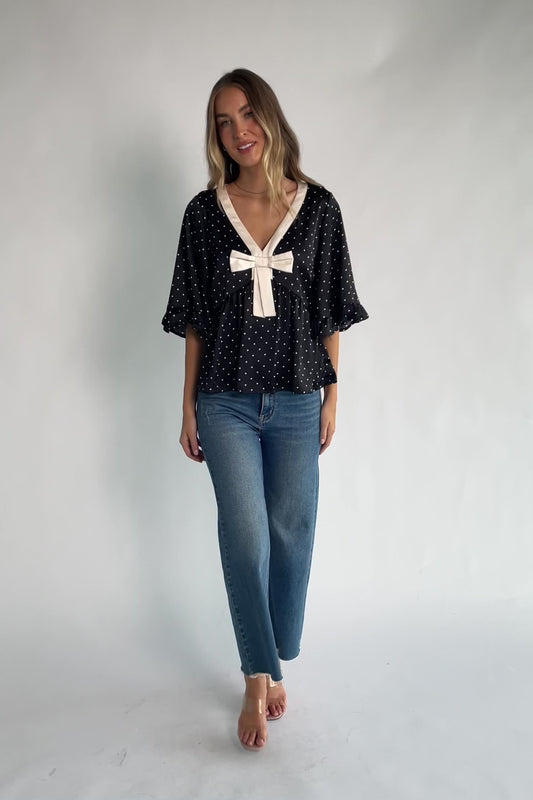 Black satin polka dot peplum top with an ivory contrast collar, decorative bow, V-neckline, and elbow-length ruffle sleeves.