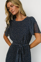 Three-quarter view of Prim Pleated Midi Dress showing taupe leaf print on deep navy chiffon fabric and the dress's flowing pleated skirt.