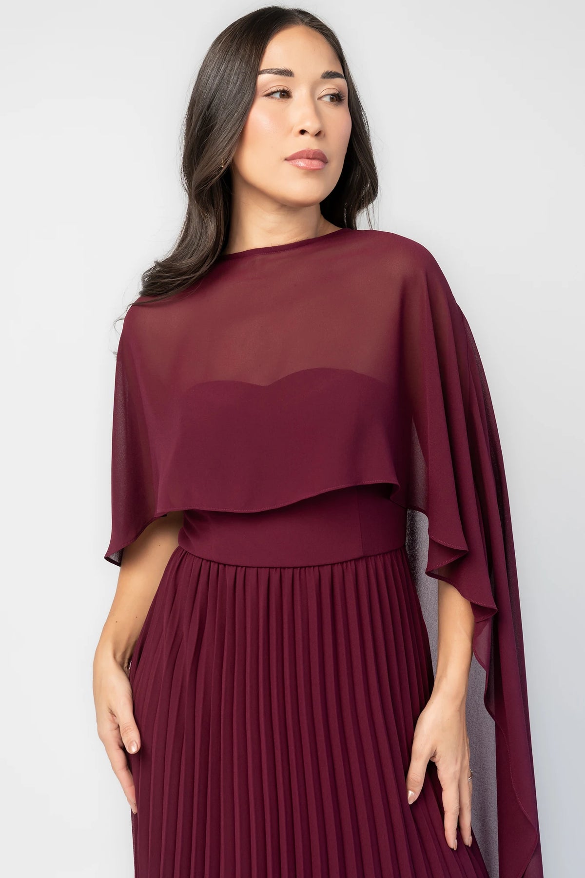 Serafina Strapless Gown in Burgundy with strapless neckline, removable sheer cape overlay, defined waist, and pleated skirt.