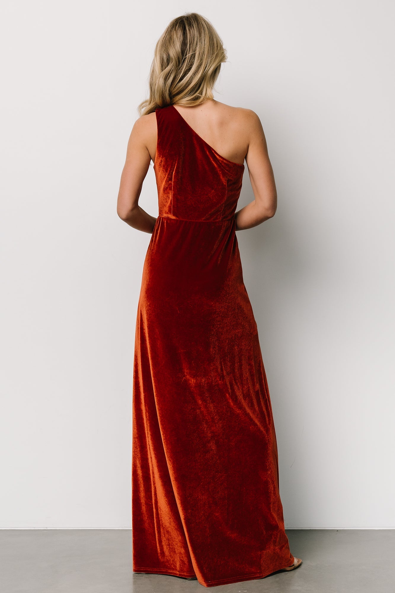 Close-up of rust velvet fabric and asymmetrical neckline of one-shoulder maxi dress, highlighting texture and stretch of the material.