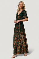 Model walking in Uma Velvet Maxi Dress in bronze and jade, showing full maxi silhouette, leg slit, and natural fabric drape.