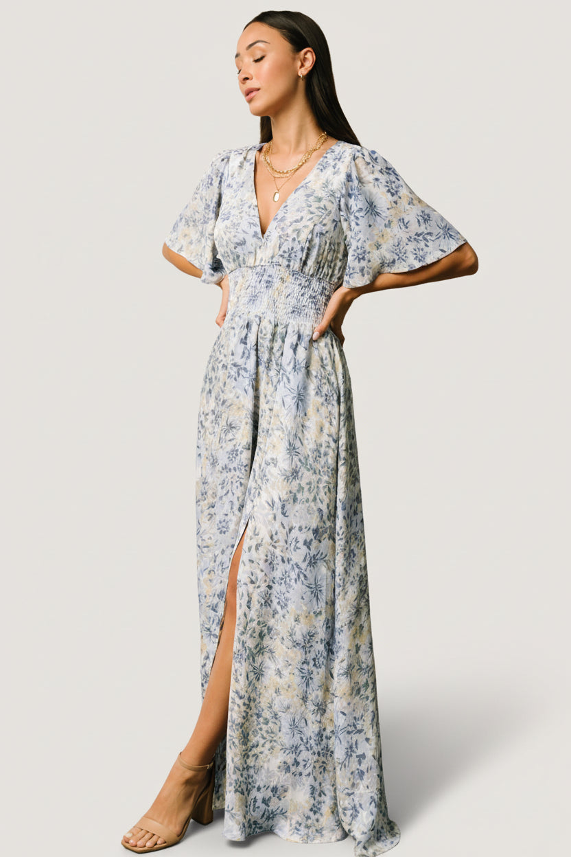Model wearing the white and blue Verona Smocked Maxi Dress showing smocked waist, deep V neckline, flowy sleeves and a left-front slit.