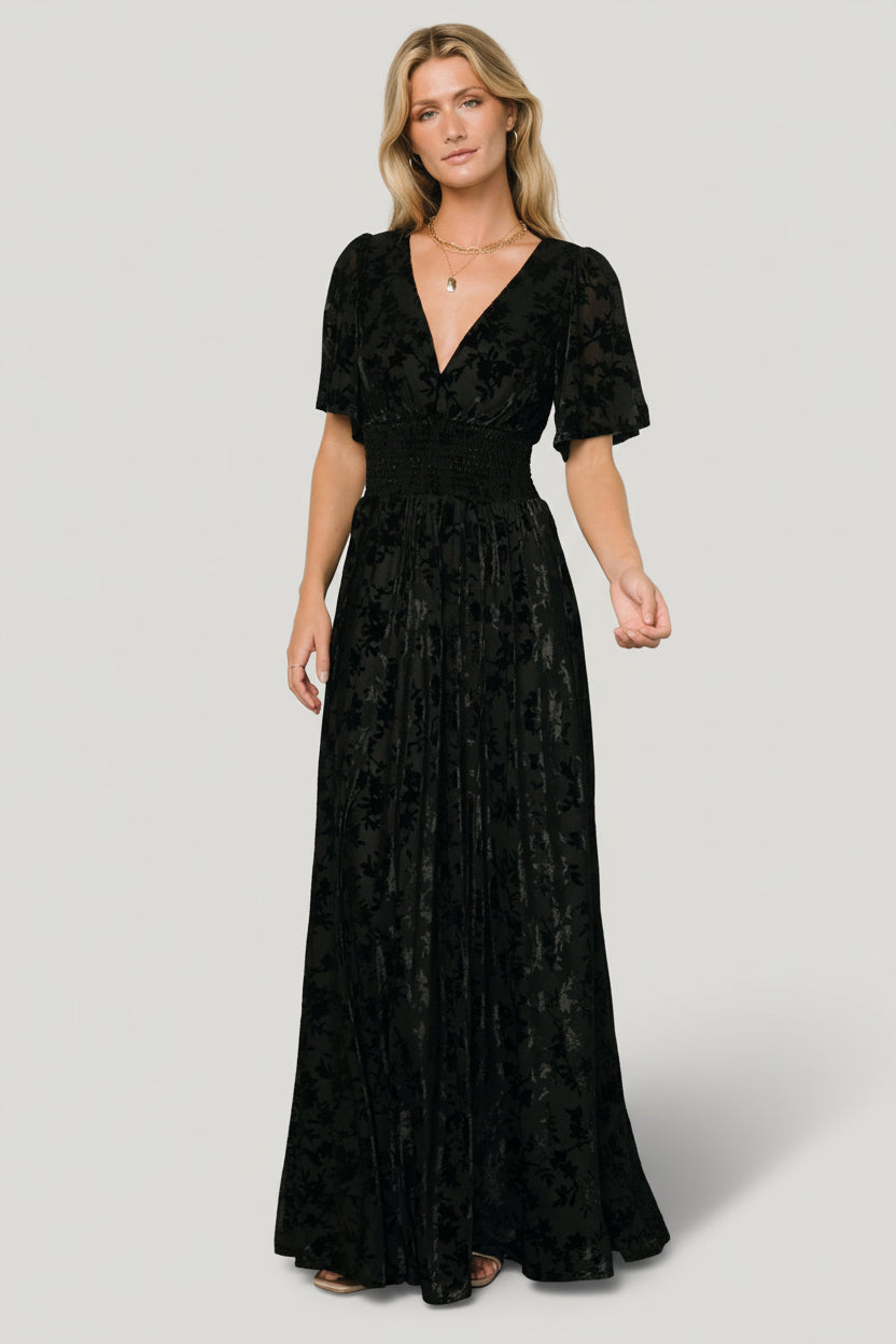 Front view of Veronica Velvet Maxi Dress in black on a model, showing deep V-neckline, smocked waist and left leg slit, short flowy sleeves.