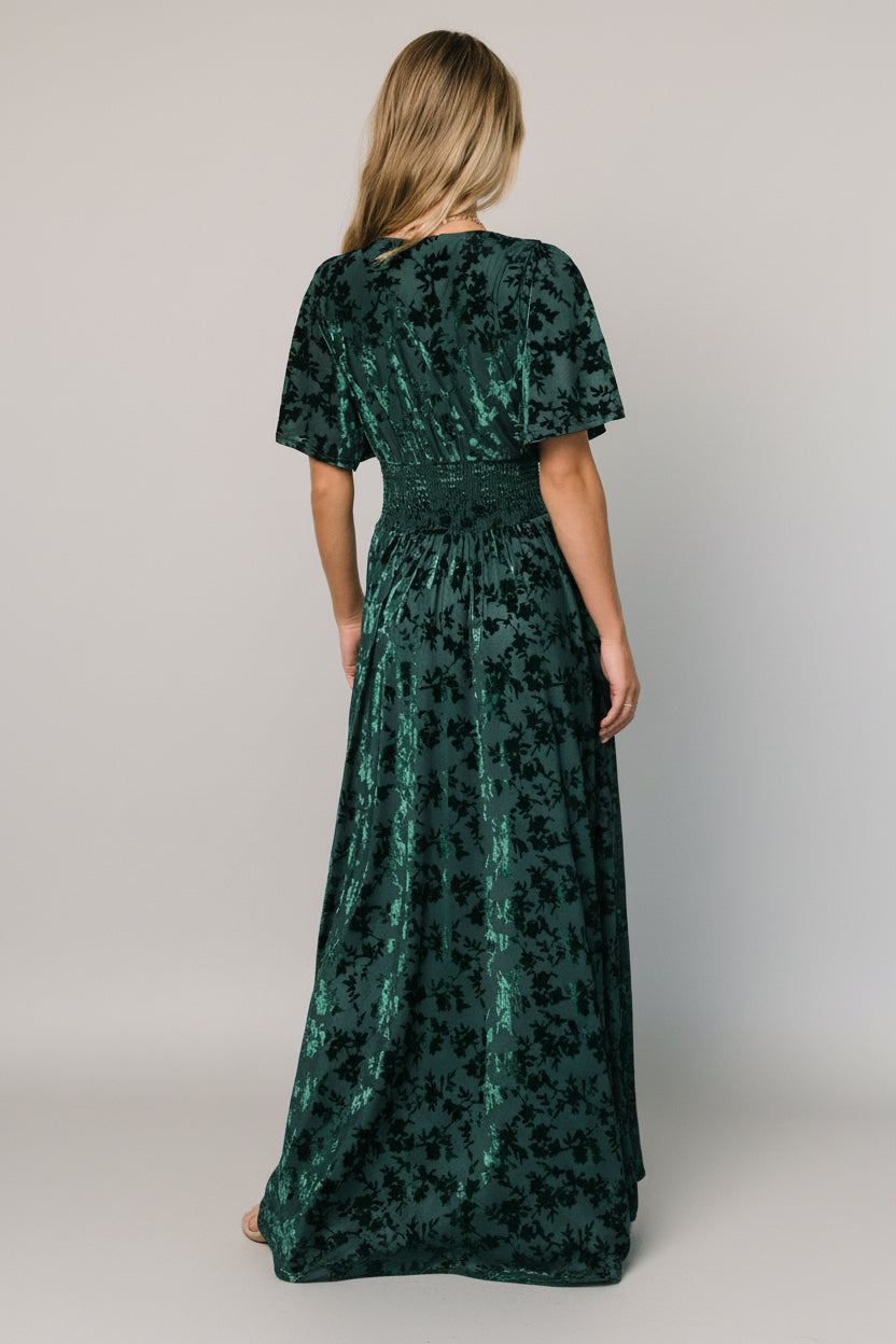 Model wearing green embossed velvet maxi dress with deep V-neck and smocked waist, showing silhouette and skirt movement.